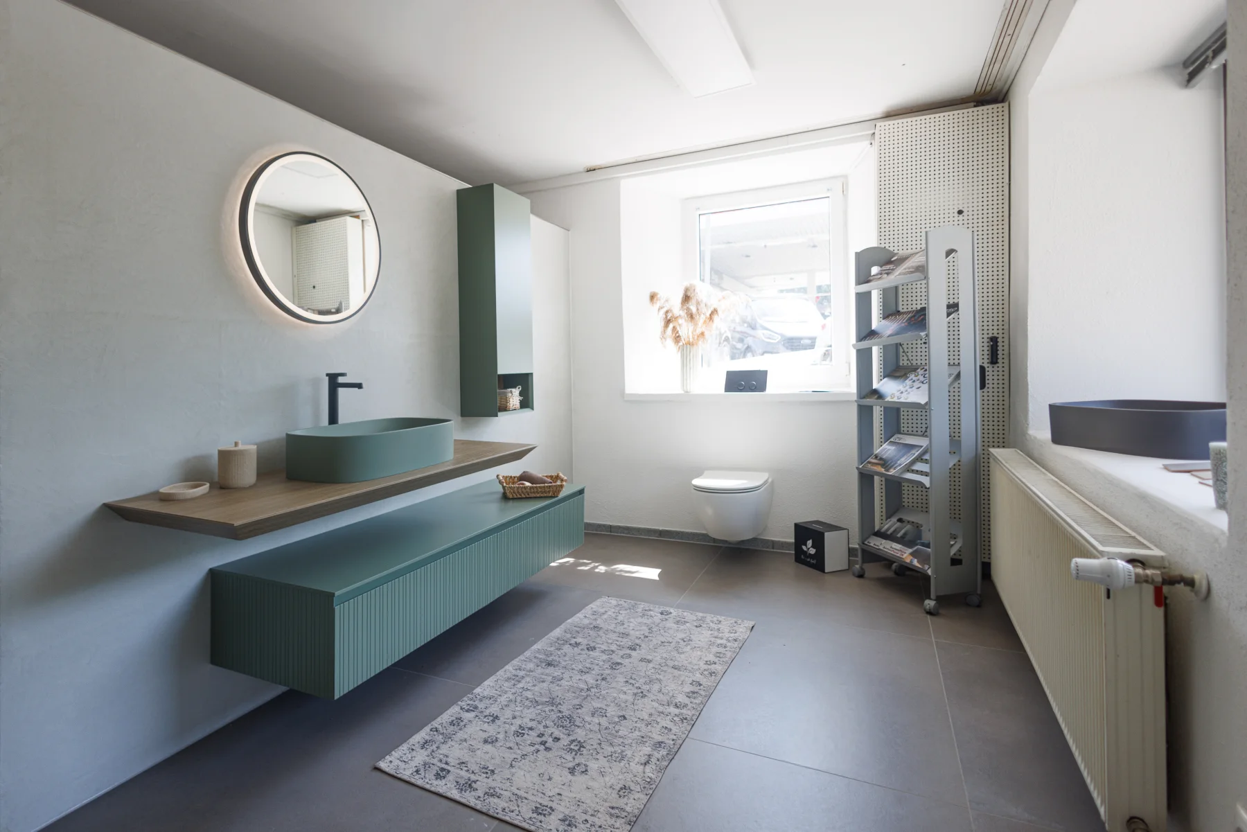 Ausstellungsbad mit mintgrünem Waschtischunterbau, Holzplatte, Rundspiegel und wandhängendem WC